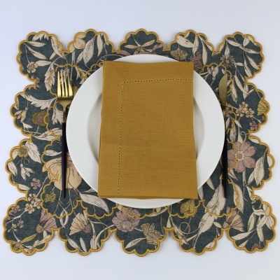 Conjunto de jantar com guardanapo castanho mostarda, prato branco, talheres dourados, e toalha de mesa floral escura com borda dourada.