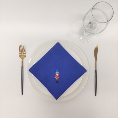 Conjunto de mesa com prato branco, guardanapo azul bordado, talheres dourados e copo de vidro
