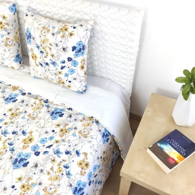 Cama com roupa de cama floral azul e amarela, mesa de cabeceira com planta e livro