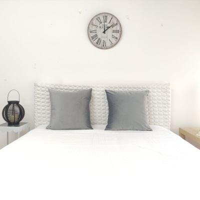 Cama branca com almofadas cinzentas, cabeceira texturada e relógio de parede