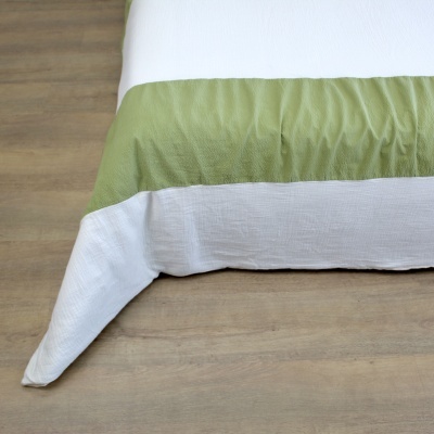 Canto de cama com lençol branco e manta verde clara sobre chão de madeira