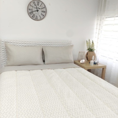 Cama com edredão bege claro e cabeceira branca, relógio de parede e mesa de cabeceira com decoração