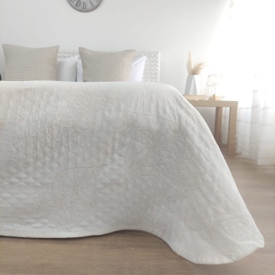 Quarto com colcha branca com padrão em relevo sobre cama, almofadas bege, relógio na parede e mesa lateral com vaso e jarra