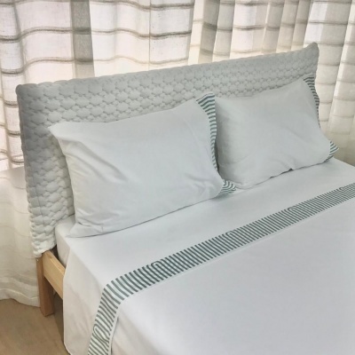 Cama com cabeceira estofada branca, almofadas brancas com bordas riscas verdes e lençol branco com faixa riscada verde