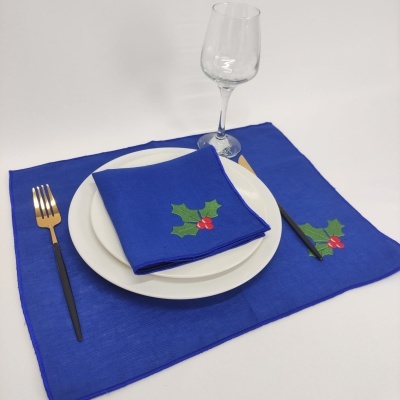 Conjunto de linha de mesa azul com folhas verdes e bagas vermelhas, prato branco, talheres pretos e dourados, e copo de vidro