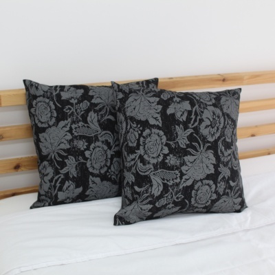 Duas almofadas com padrão floral cinzento sobre fundo preto em cama com roupa branca e cabeceira de madeira.