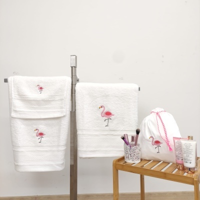 Toalhas brancas com bordado de flamingo rosa e acessórios de banho sobre banco de madeira