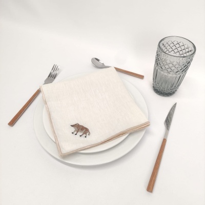 Talheres com cabo em madeira clara, guardanapo branco com borda bege e raposa bordada, prato branco e copo transparente com textura