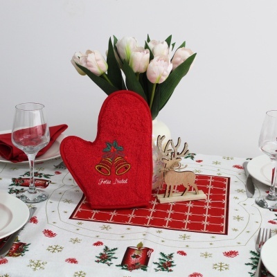Luvas de cozinha vermelhas natalícias sobre toalha decorada com flores, vaso branco com flores rosa e figura de rena