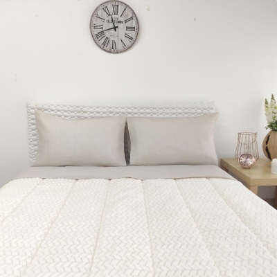 Cama com edredão branco, cabeceira trançada, almofadas bege, relógio de parede e mesa de cabeceira com flores