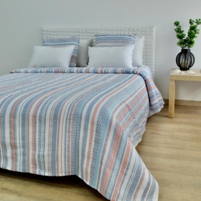 Cama com colcha riscada azul, branco e rosa, almofadas combinando, mesa ao lado com vaso preto e planta