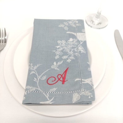 Guardanapo azul cinza com padrão floral branco e letra A vermelha bordada sobre prato branco