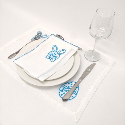 Toalhete e guardanapo de tecido branco bordados a azul com desenhos de coelho e círculo floral, prato, talheres e copo