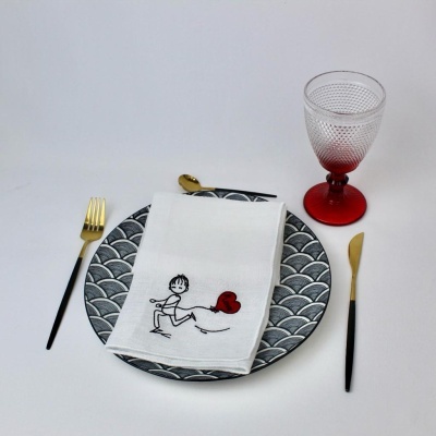 Conjunto de mesa com prato cinzento e preto, guardanapo branco com desenho, talheres pretos e dourados e copo com pé vermelho