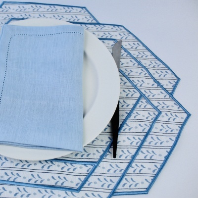 Jogo americano branco com padrão azul, guardanapo azul claro sobre prato branco e talheres pretos.