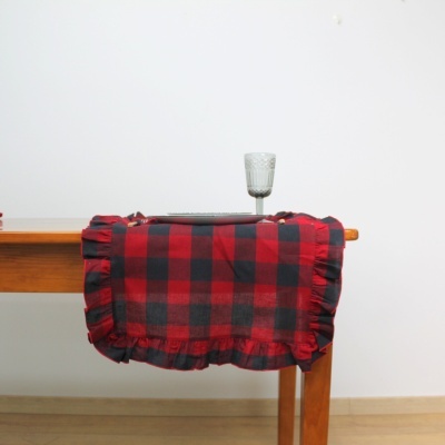Manta xadrez vermelha e preta com barra franzida pendurada sobre uma mesa de madeira clara com taça de vidro em cima.