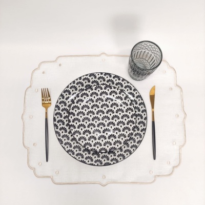 Mesa posta com prato decorado preto e branco, talheres dourados e pretos, copo transparente e individual branco bordado