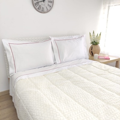 Cama com colcha creme texturizada, fronhas brancas com borda vermelha e mesa de cabeceira com vaso e livros.