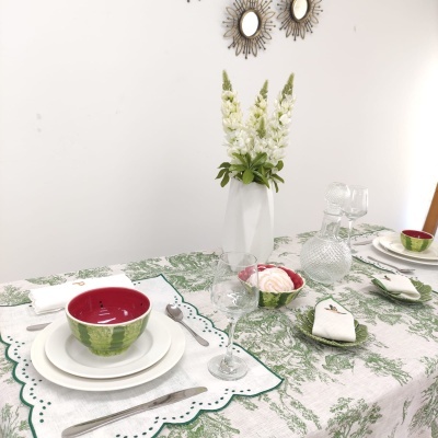 Mesa de jantar com toalha verde, pratos brancos, tigelas vermelhas e brancas, copos e talheres, vaso branco com flores, parede branca com dois espelhos redondos
