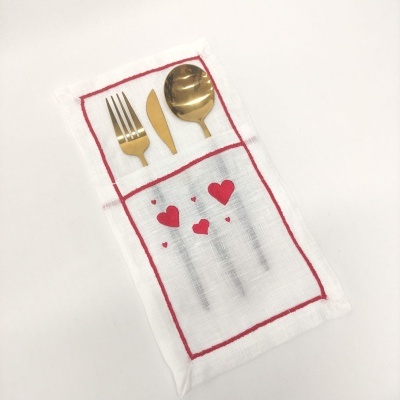 Talheres dourados sobre toalha branca com bordado de corações vermelhos e linhas vermelhas formando bolsos