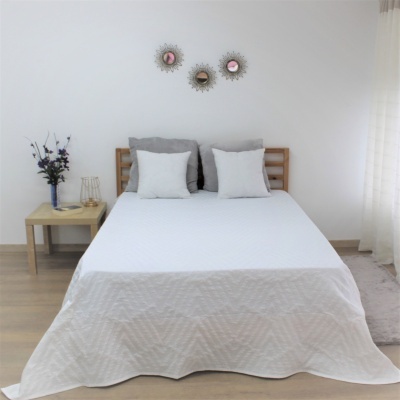 Cama de madeira com colcha branca e almofadas cinza claras numa divisão branca com mesa de cabeceira e vaso de flores