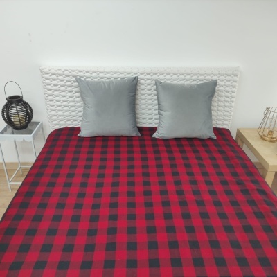 Cama com colcha xadrez vermelha e preta, duas almofadas cinzentas, cabeceira branca texturizada e mesas laterais com candeeiros