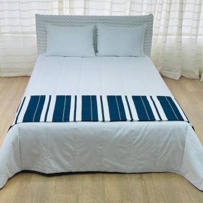 Cama com lençóis brancos, edredão branco com lados pretos e lençol decorativo azul listrado, em chão de madeira clara e cortinas brancas ao fundo