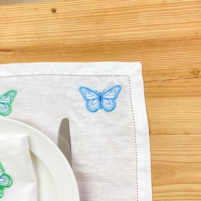 Guardanapo branco tecido com bordado de borboletas azul e verde junto a prato e faca sobre madeira