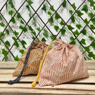 Dois sacos de tecido com padrão branco e cordões coloridos sobre superfície de madeira com treliça e folhas verdes ao fundo