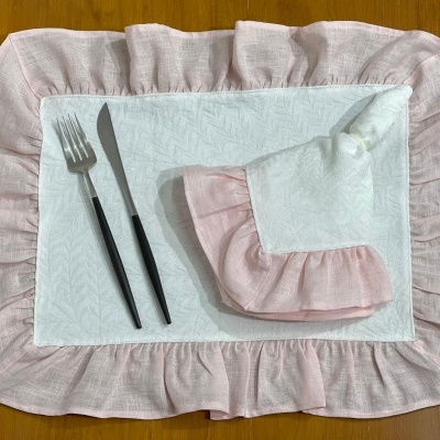 Jogo de mesa com individual branco e rosa, guardanapo com argola e talheres sobre mesa de madeira.