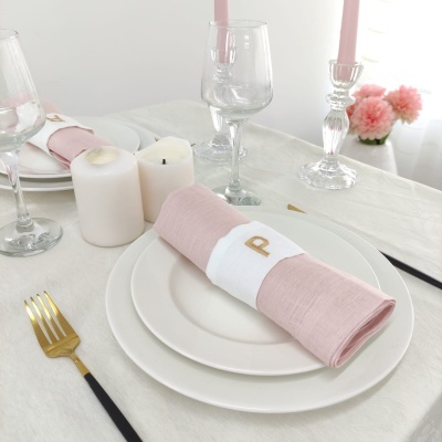 Mesa posta com prato branco, guardanapo rosa com argola branca com letra 'P', talheres dourados e pretos, copos de vidro e velas brancas.