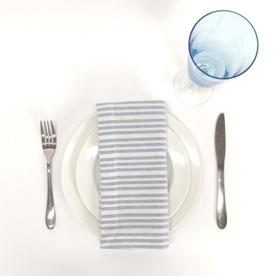 Conjunto de mesa com pratos brancos, talheres de aço, guardanapo riscado e copo azul
