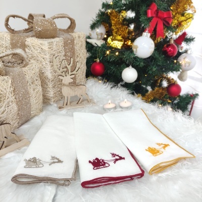 Três toalhas bordadas com temas de trenó e rena junto a árvore de Natal e presentes decorativos.