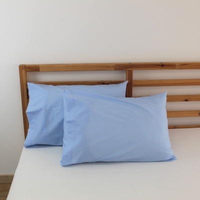 Duas almofadas com fronhas azuis claras numa cama de madeira clara com colchão branco e parede branca ao fundo.