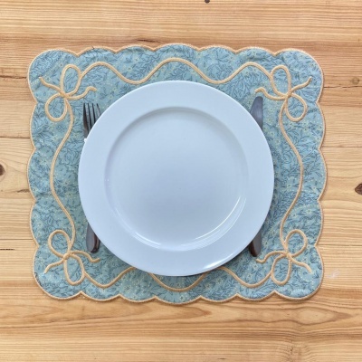 Lugar americano azul claro com padrão floral e bordas onduladas, prato branco, talheres e fundo de madeira