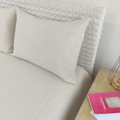 Almofadas e lençol bege claro com padrão, cabeceira branca texturizada, mesa de cabeceira com livros e vela