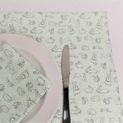 Jogo americano e guardanapo branco com desenhos de coelhos e esquilos cinza, prato branco e faca preta
