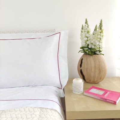 Almofada e cama branca com bordado vermelho, vaso de madeira com flores, vela e livro rosa na mesa
