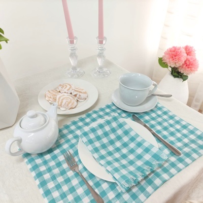 Conjunto de chá branco com bule, chávena com pires e colher junto a guardanapo azul xadrez, prato com bolachas, castiçais com velas cor-de-rosa e vaso de flores cor-de-rosa.