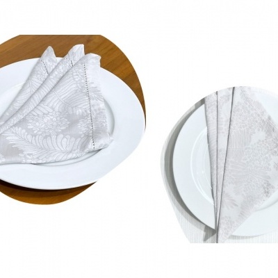 Dois conjuntos de pratos brancos com guardanapos de tecido cinza claro com padrão floral dobrados sobre eles em superfícies diferentes