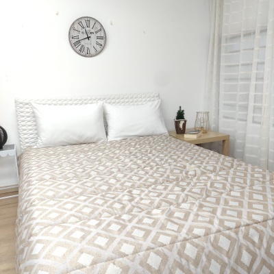 Quarto com cama de casal, roupa de cama bege com padrão geométrico branco, cabeceira branca, relógio de parede e mesa de cabeceira com planta.