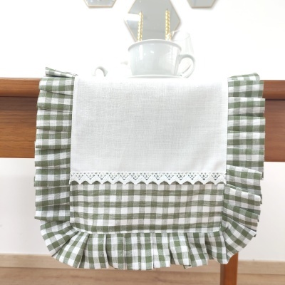Toalha decorativa xadrez verde e branco com área branca e detalhes bordados pendurada numa mesa de madeira clara