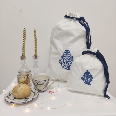 Dois sacos de tecido branco com padrão azul, prato e chávena com pão, candeeiros e luzes numa superfície branca