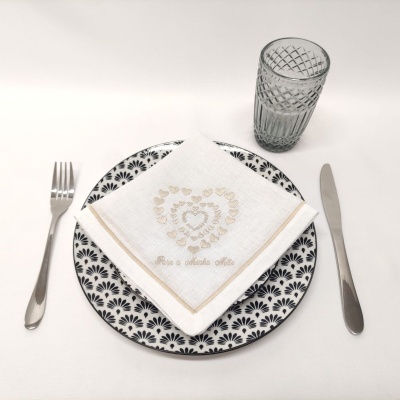 Conjunto de jantar com prato branco com padrão preto, guardanapo branco bordado, copo de vidro e talheres sobre toalha branca