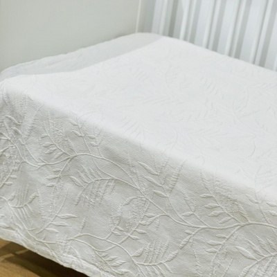 Colcha branca texturizada com padrão de folhas numa cama branca de ripas, chão de madeira