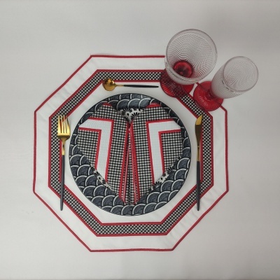 Conjunto de mesa com prato, talheres dourados, guardanapo, copos vermelhos e placemats octogonais em tecido branco e vermelho