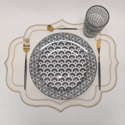 Conjunto de louça com prato branco e padrão preto, talheres dourados e copo transparente sobre jogo americano branco