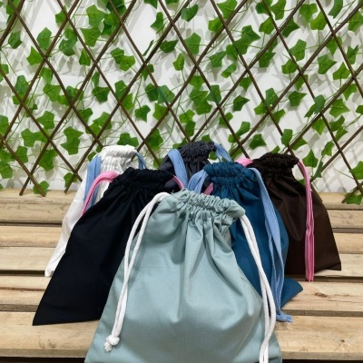 Sete bolsas de tecido tipo saco coloridas com cordões, sobre madeira e treliça com folhas verdes ao fundo