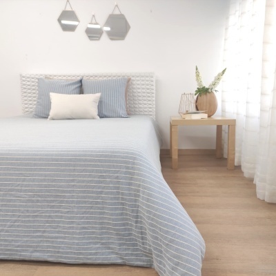 Quarto com cama de tecido branco texturizado, edredão azul com riscas brancas, mesa de madeira com vaso e flores, três espelhos geométricos na parede