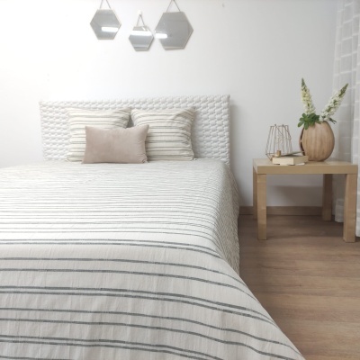 Cama com roupa de cama riscada cinzenta e bege, cabeceira branca e mesa de madeira com vaso e flores